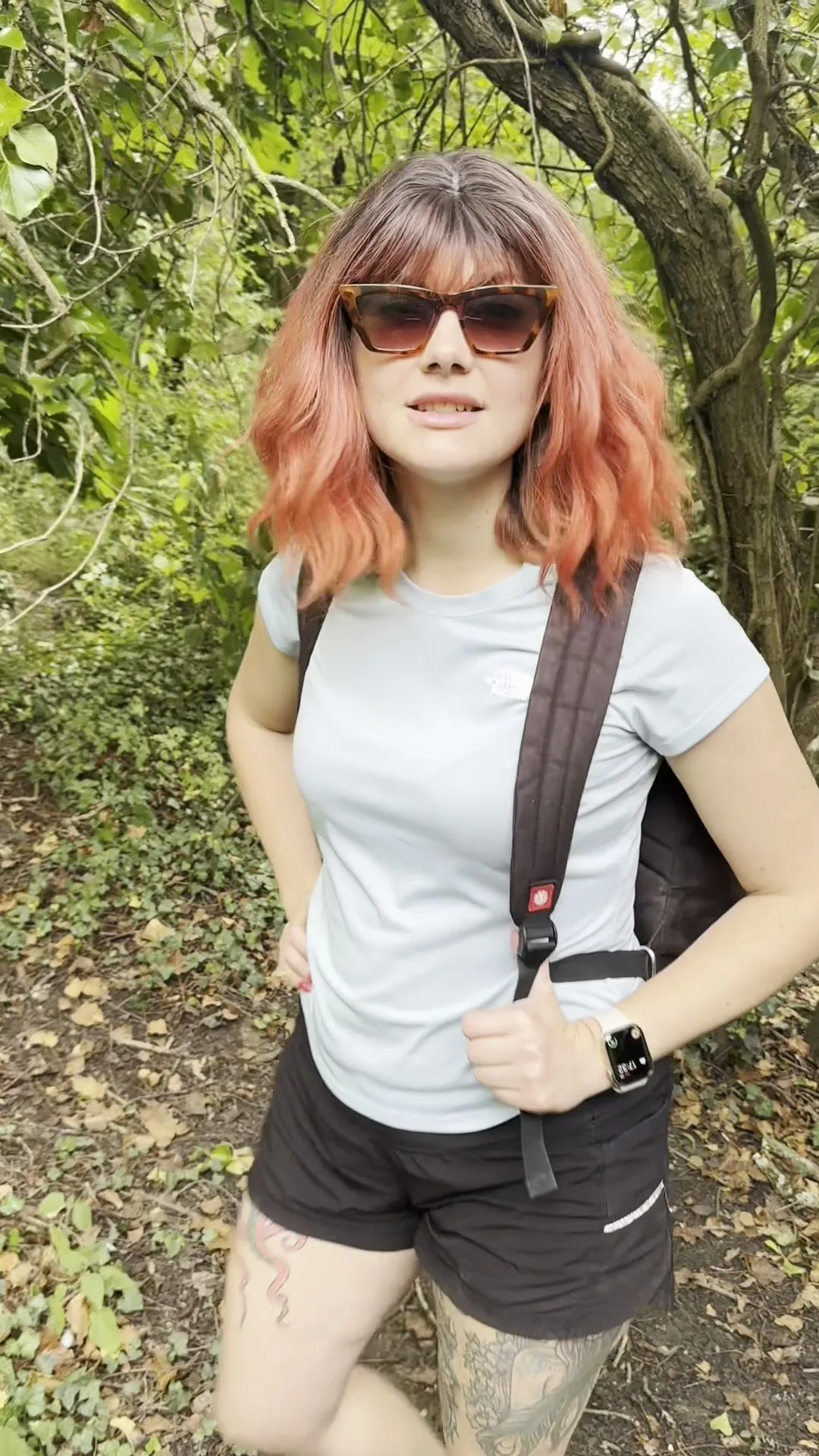 Redheaded girl with leg tattoos walks and poses in forest with backpac
